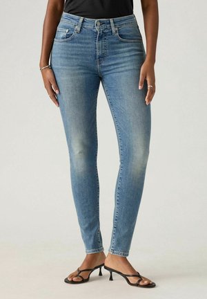 Kvinde iført lyseblå skinny jeans, sort ærmeløs top, sorte hæle sandaler samt sølvringe og armbånd, står mod en ensfarvet baggrund.