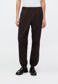 Σκούρο καφέ φόρμες με ελαστική μέση και σούρες στα μανσέτα, κατασκευασμένες από μαλακό ύφασμα. Συνδυασμένες με λαμπερά μαύρα loafers.