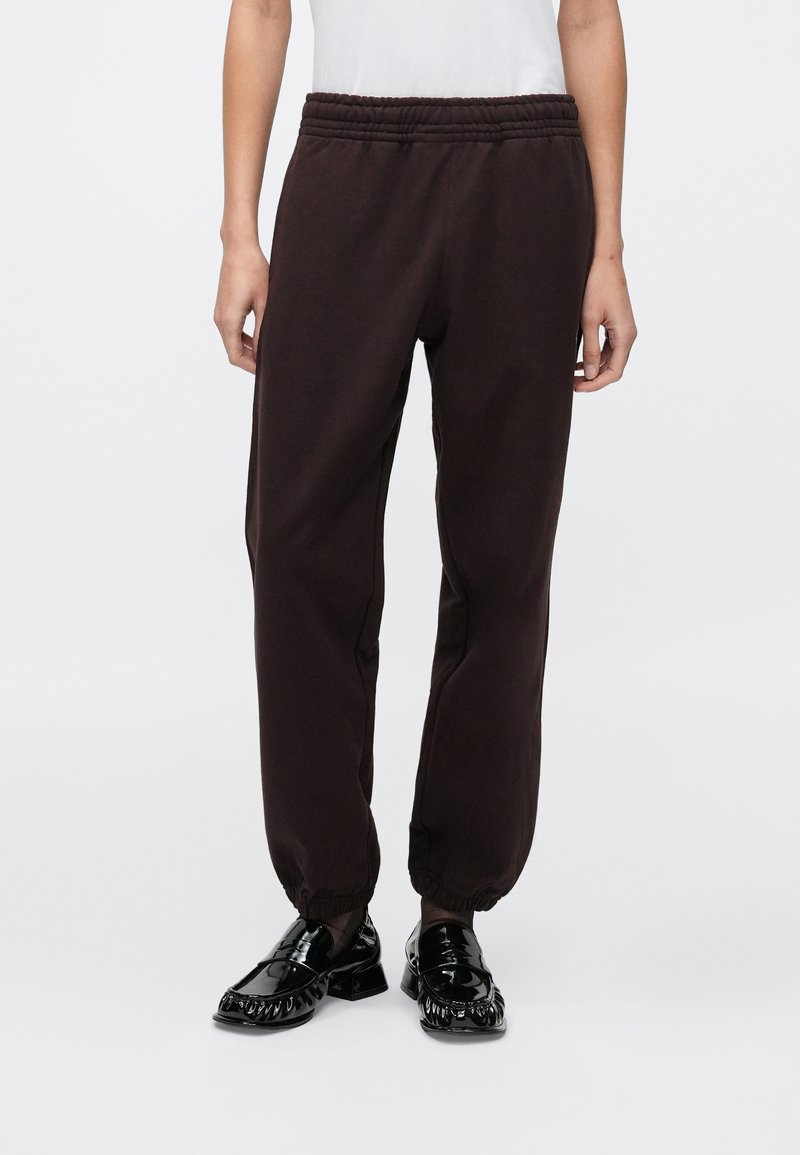 Σκούρο καφέ φόρμες με ελαστική μέση και σούρες στα μανσέτα, κατασκευασμένες από μαλακό ύφασμα. Συνδυασμένες με λαμπερά μαύρα loafers.