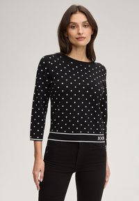 Μαύρο πουλόβερ με λευκές πουά, μανίκια τριών τετάρτων και τελείωμα με λευκή τρέσα που φέρει λεπτομέρεια με λογότυπο στο κάτω μέρος.