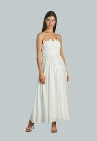 Witte maxi-jurk met een bodice van bloemenkant, spaghettibandjes en een vloeiende rok; gladde textuur. Model draagt sandaaltjes met bandjes, lichte achtergrond.