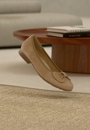 Beiger Wildleder-Ballerina mit Schleifendetail, die über einem strukturierten Teppich, einem runden Holzcouchtisch und gestapelten Büchern in einem gemütlichen Wohnzimmer schwebt.