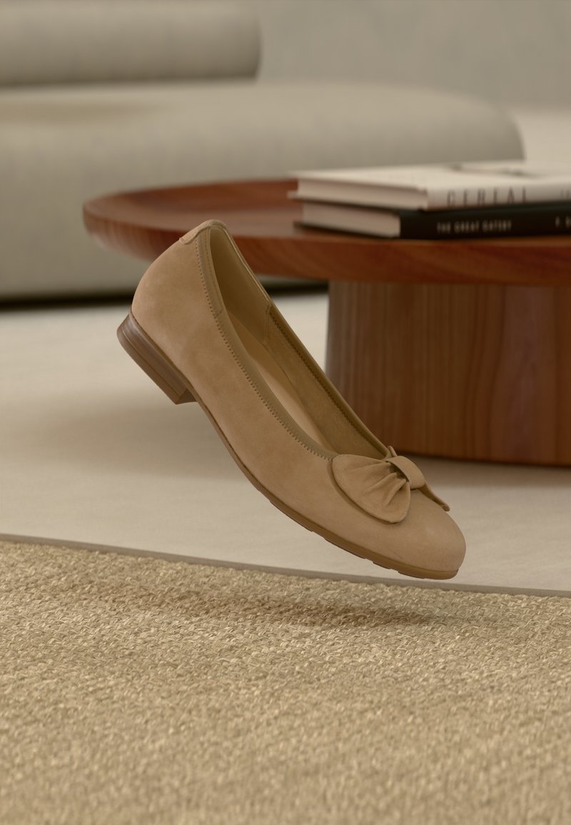 Μπεζ σουέντ μπαλαρίνες με φιόγκο που αιωρούνται πάνω από ένα υφασμάτινο χαλί, στρογγυλό ξύλινο τραπεζάκι σαλονιού και στοιβαγμένα βιβλία σε ένα απαλό περιβάλλον καθιστικού.