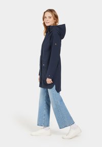 Marineblauer wasserdichter Parka mit Kapuze, zwei Fronttaschen, Druckknöpfen und langem Schnitt, kombiniert mit weit geschnittenen hellblauen Jeans und weißen Sneakern.