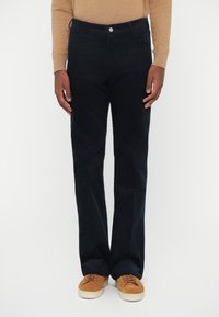 Tumši jūras zilā korduroja bikses ar taisnu piegriezumu, ar klasisku pogu jostu, priekšējām kabatām un mīkstu tekstūru. Valkātas ar brūnām tenisa kurpēm.