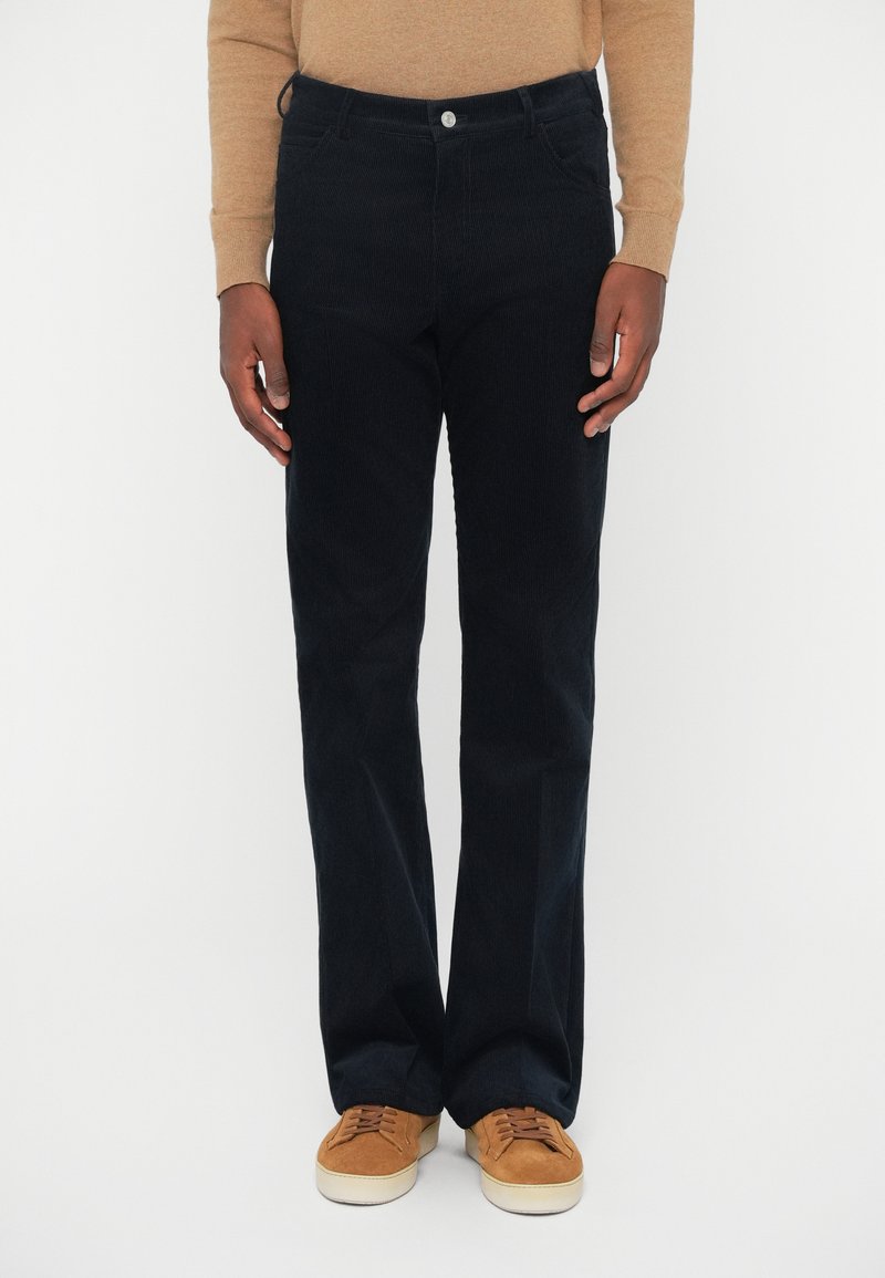 Dunkelblaue Cordhosen mit geradem Schnitt, ausgestattet mit einem klassischen Knopfverschluss, Fronttaschen und einer weichen Textur. Getragen mit braunen Sneakers.