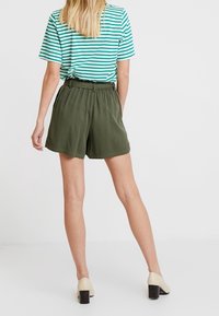 Pantaloni verde in tessuto strutturato con cintura elastica, abbinati a una T-shirt a righe verdi e bianche. Scarpe beige con tacco a blocco.