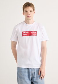Balta kokvilnas T-krekls ar taisnstūra formas sarkanu grafiku, kurā spēlē uzraksts "CALVIN NEW KLEIN YORK" treknajā fontā. Brīvs piegriezums, īsas piedurknes, minimālistisks dizains.