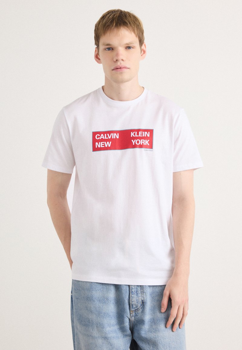 Balta kokvilnas T-krekls ar taisnstūra formas sarkanu grafiku, kurā spēlē uzraksts "CALVIN NEW KLEIN YORK" treknajā fontā. Brīvs piegriezums, īsas piedurknes, minimālistisks dizains.