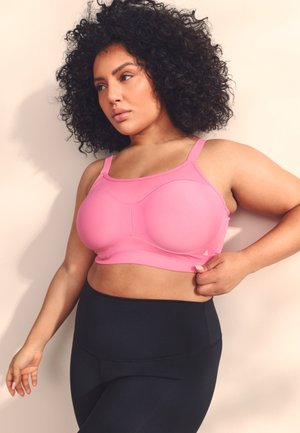 Žena s kučeravými vlasmi, oblečená v ružovom športovom podprsenke a čiernych legínach, upravuje ramienko podprsenky, stojí na svetlom pozadí.