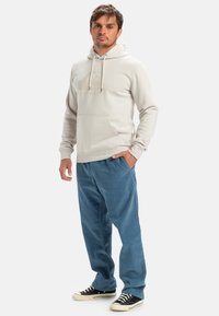 Hoodie beige dengan saku depan dan tudung tali, dipadukan dengan celana korduroi biru dan sepatu sneakers hitam, yang memiliki sol karet putih.