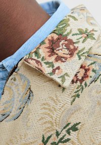 Jas van stof met bloemenpatroon, met een beige basis en roze en groene accenten. De kraag is van denim in een lichte blauwe kleur.