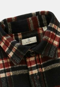 Chemise en flanelle à carreaux noire, rouge et crème avec un col boutonné et une étiquette de marque visible à l'intérieur du col.