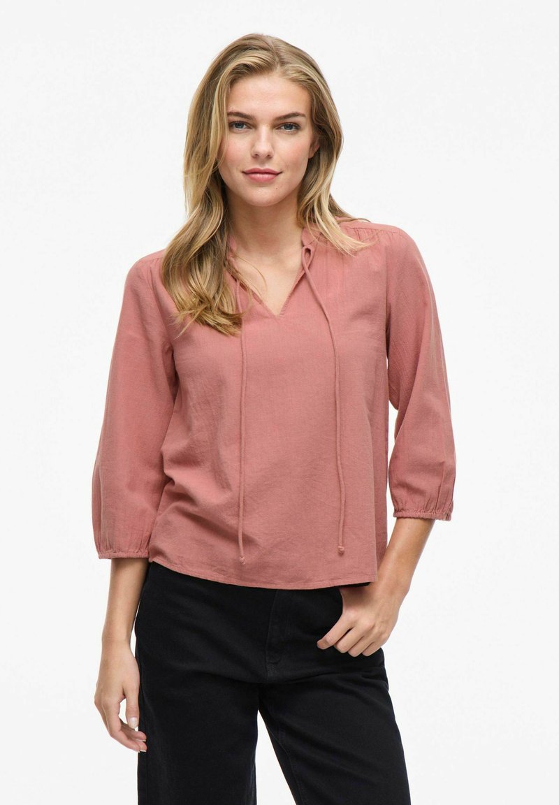 Dammig rosa pulloverblus med V-ringning och justerbar dragsko. Har 3/4-längd ärmar med samlade muddar och mjuk textur.