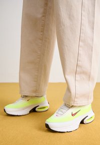 Neongröna och vita Nike-sneakers med strukturerad överdel, svarta detaljer och luftdämpning, ihopparade med beigefärgade vida byxor.