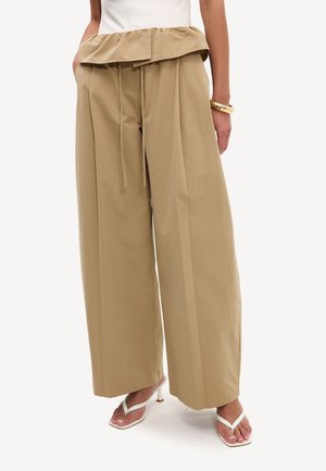 Pantalon beige à jambes larges avec une ceinture haute pliée et un cordon, porté avec des sandales blanches à talons.