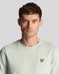 Homme avec des cheveux bruns courts portant un t-shirt vert clair avec un petit logo d'oiseau jaune sur la poitrine, se tenant face à un fond gris.