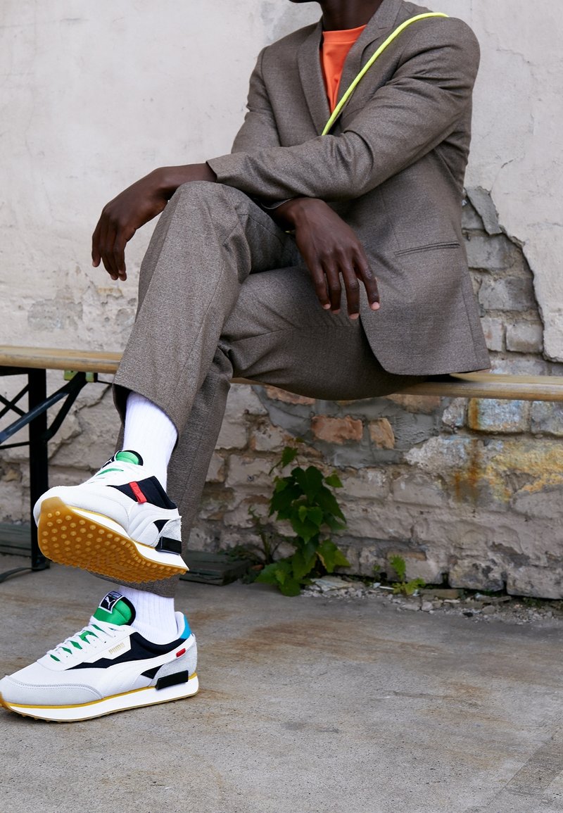 Homme portant un costume gris, une chemise orange, des chaussettes blanches et des baskets blanches, assis de façon décontractée sur un banc contre un mur en briques usé.