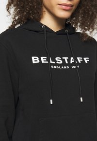 Svart hoodie i mjukt tyg, med en vit tryckt logotyp "BELSTAFF ENGLAND 1914" och en framficka. Dragkedjorna har silverfärgade ändar.