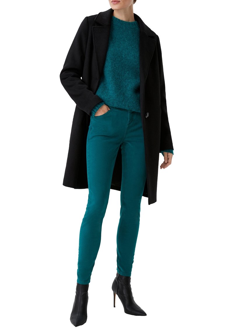 Abrigo negro de lana, suéter esponjoso teal y jeans ajustados teal, combinados con botas de tacón negro. El atuendo presenta una textura superpuesta.