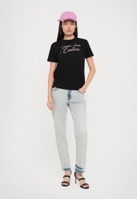 Juodas medvilninis marškinėlis su "Versace Jeans Couture" tekstu rožinės spalvos. Derinama su šviesiai mėlyniais džinsais ir juodomis aukšto kulno sandalais, papildyta rožiniu kepurėliu.