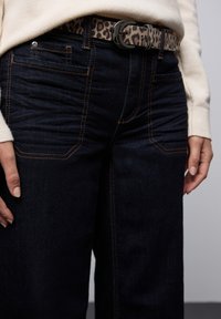Jean en denim noir avec une coupe droite, featuring des coutures orange, des poches avant et une ceinture à imprimé léopard avec une boucle noire.