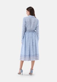 Light blue dress with long sleeves, patterned fabric, smocked waist, and a decorative hem. Paired with light blue high-heeled sandals.