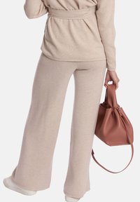 Pull en tricot beige avec ceinture et pantalon large assorti ; texture lisse. Le modèle tient un petit sac seau en cuir marron avec une bandoulière.