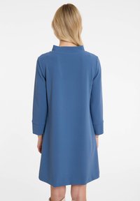 Robe bleue jusqu'aux genoux avec un col haut et des manches trois-quarts. Texture lisse, design simple et fentes latérales pour faciliter le mouvement.