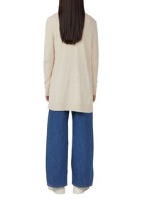 Donna con lunghi capelli scuri, che indossa un maglione beige a maniche lunghe, jeans larghi blu e sneakers bianche, di spalle su sfondo bianco.