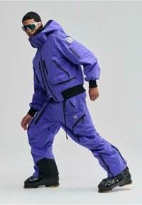 Lila vízálló síruházat, több fekete cipzárral, hosszú kapucnival és eltérő fekete alsó kiegészítőkkel; fekete sícipővel viselve.