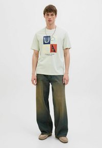Lys grøn t-shirt med et firkantet grafik, der viser forskellige former og farver; parret med brede, blege denim jeans og lysebrune sko.