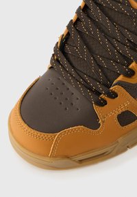 Sapatilha castanha e bege com couro texturizado e malha. Apresenta atacadores escuros, perfurações na ponta e uma sola de borracha.