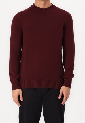 Mann trägt einen schlichten, bordeauxroten, langärmeligen Strickpullover mit gerippten Bündchen, Saum und Rundhalsausschnitt, kombiniert mit einer schwarzen Hose.