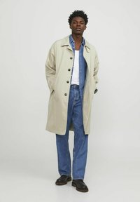 Beige regnrock med svarta knappar, bärs över en denimskjorta och blå jeans; svarta skor; strukturerad tyg; lös passform; avslappnad stil.