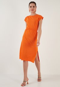 Oranges Midi-Kleid mit kurzen Ärmeln, elastischem Bund, seitlichem Schlitz und weichem Baumwollmaterial. Kombiniert mit Riemchen-Heelsandalen.