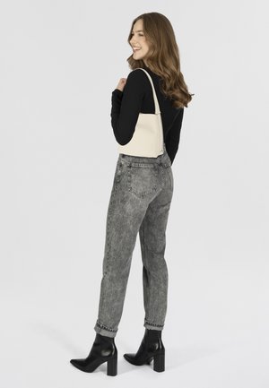 Sac à main en cuir beige avec un design minimaliste. Modèle portant un pull noir et un jean taille haute gris, associé à des bottes à talons noires.