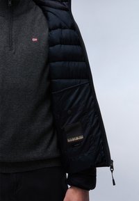 Černá puffer bunda uvnitř s tmavě šedým svetrem s malou vlajkovou nášivkou. Obsahuje kapsu na zip s logem značky.