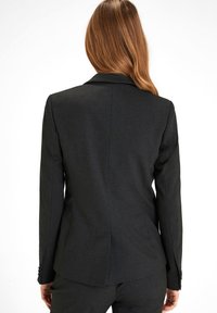 Blazer nero su misura con uno spacco posteriore, revers a punta e maniche lunghe con dettagli bottonati. Tessuto dalla consistenza liscia.