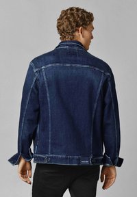 Giacca in denim blu scuro con taglio classico, cuciture visibili e polsini con bottoni. Presenta il marchio Red Bull Racing sull'orlo.