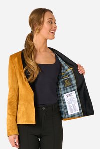 Dviejų spalvų švarkas su garstyčių spalvos zomšiniu išoriniu paviršiumi ir juoda vidine dalimi. Turi languotą pamušalą su atspausdintu tekstu bei vieną sagą užsegimui.