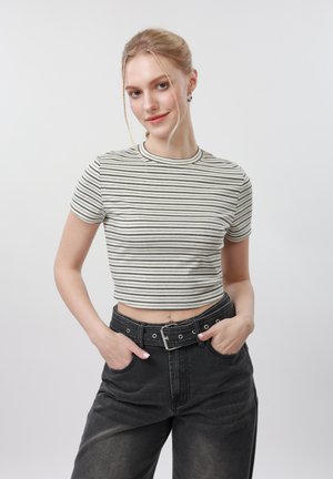 Lyshåret kvinde med håret sat op i en knold, iført en stribet, kort t-shirt og sorte højttaljede jeans med bælte, stående med hænderne i lommerne.