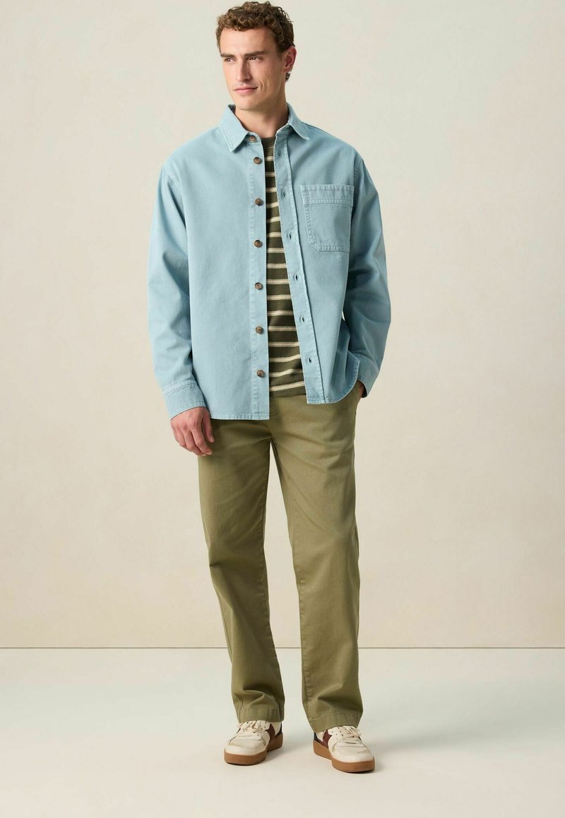 Chemise bleue claire boutonnée sur un t-shirt à rayures vertes, assortie à un pantalon olive et des baskets beiges. Tissu lisse, coupe décontractée.