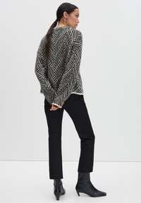 Frau mit langen, dunklen Haaren in einem tiefen Pferdeschwanz, die einen schwarz-weißen Zickzack-Pullover, schwarze Hosen und schwarze Absatzstiefel trägt, steht mit Händen hinter dem Rücken.