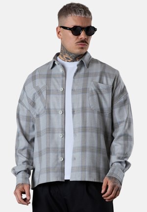 Mann mit runden schwarzen Sonnenbrillen, weißem T-Shirt und grauem karierter Hemd, mit Tätowierungen am Hals und an den Händen.