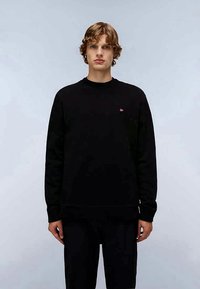 Jovem com cabelo encaracolado, vestindo uma camiseta preta de manga longa básica e calças pretas, de pé contra um fundo cinza-claro.