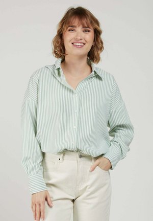 Femme souriante aux cheveux courts et ondulés porte une chemise rayée vert clair et blanc boutonnée et un pantalon taille haute écru, posant avec une main dans la poche.