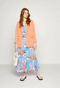 Blazer coral sobre um vestido floral azul com detalhes em rosa e amarelo, combinado com chinelos brancos e uma mala creme.