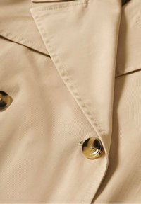 Tissu de trench beige avec une texture lisse, présentant un gros bouton brun brillant et un détail de col plié sur le côté gauche.