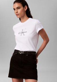 Biele bavlnené tričko s vyhrnutými rukávmi a sivým logom Calvin Klein, doplnené o čiernu rifľovú sukňu so strieborným opaskom s prackou.